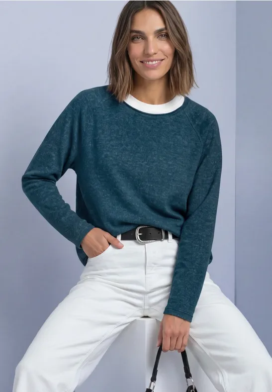 Vrouw zittend voor lichte achtergrond, blauwgroene trui, witte broek en glimlach.