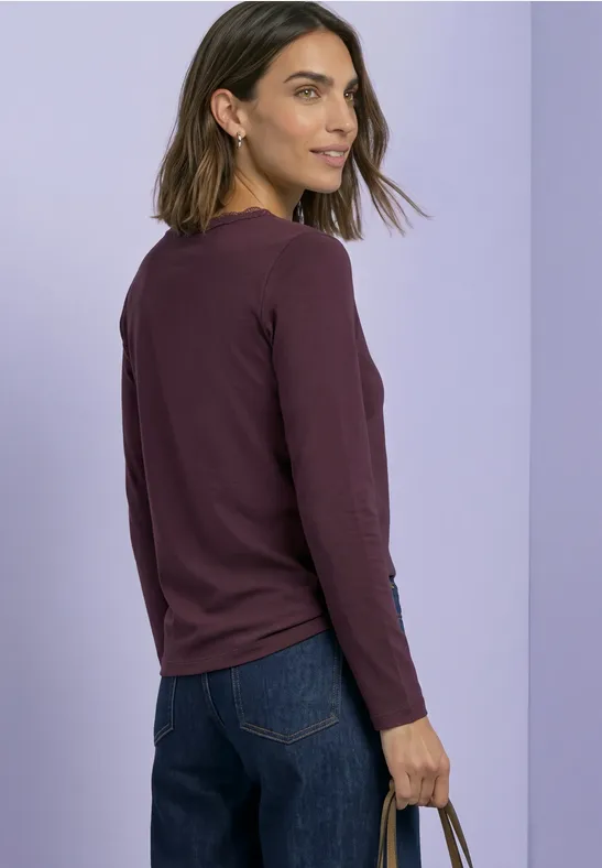 Vrouw in bordeaux longsleeve en spijkerbroek, zijaanzicht van achter, met handtas tegen lichte achtergrond