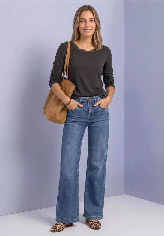 Frau steht vor neutralem Hintergrund, trägt schwarzen Pullover, weite Jeans, Sandalen und eine beige Schultertasche.
