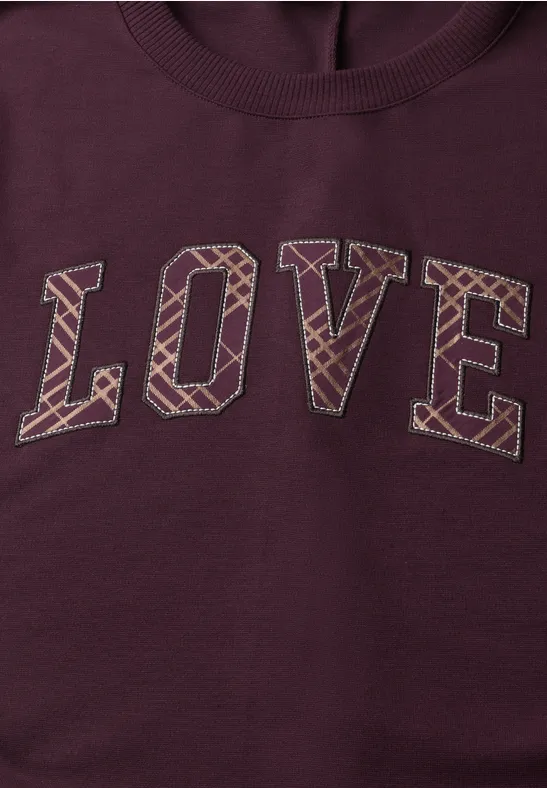 Bordeauxfarbener Pullover mit dem Schriftzug LOVE in großen Blockbuchstaben und geometrischem Muster.