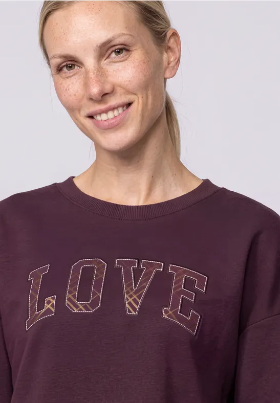 Eine lächelnde Frau mit hellblondem Haar im Pferdeschwanz, trägt ein violettes Sweatshirt mit dem Schriftzug 'LOVE'.