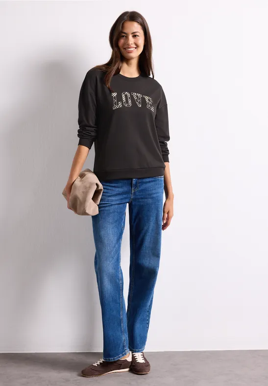 Junge Frau im schwarzen Pullover mit LOVE-Schriftzug und blauen Jeans, lächelt und hält einen beigen Hut.