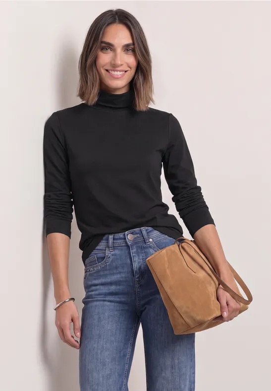 Eine lächelnde Frau in schwarzem Rollkragenpullover und Jeans, hält eine braune Handtasche.