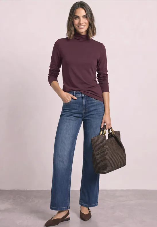 Vrouw in bordeaux coltrui en blauwe jeans, met een bruine tas en zwarte ballerina's.