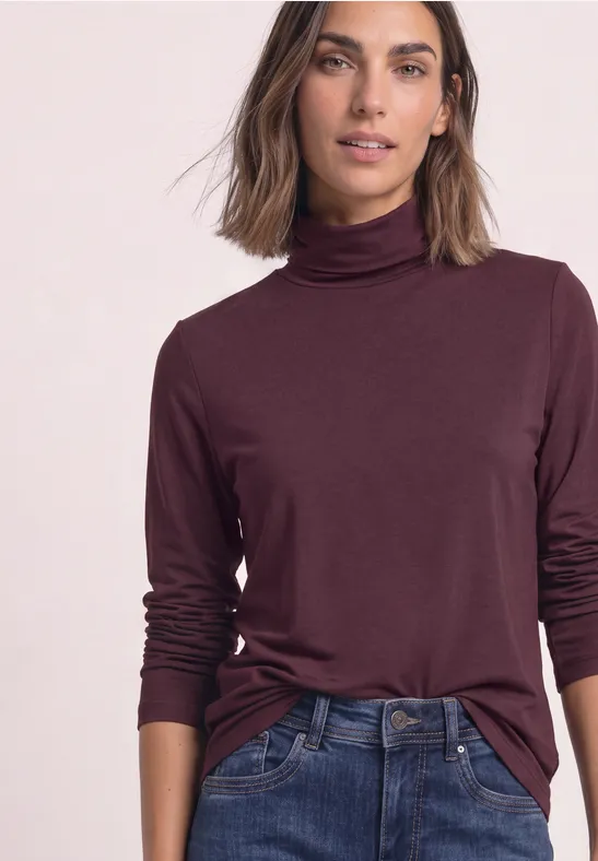 Vrouw in bordeaux coltrui en blauwe spijkerbroek staat casual.
