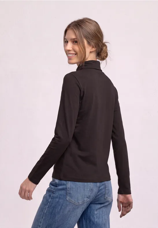 Junge Frau in schwarzem Langarmshirt und Jeans blickt lächelnd über die Schulter.