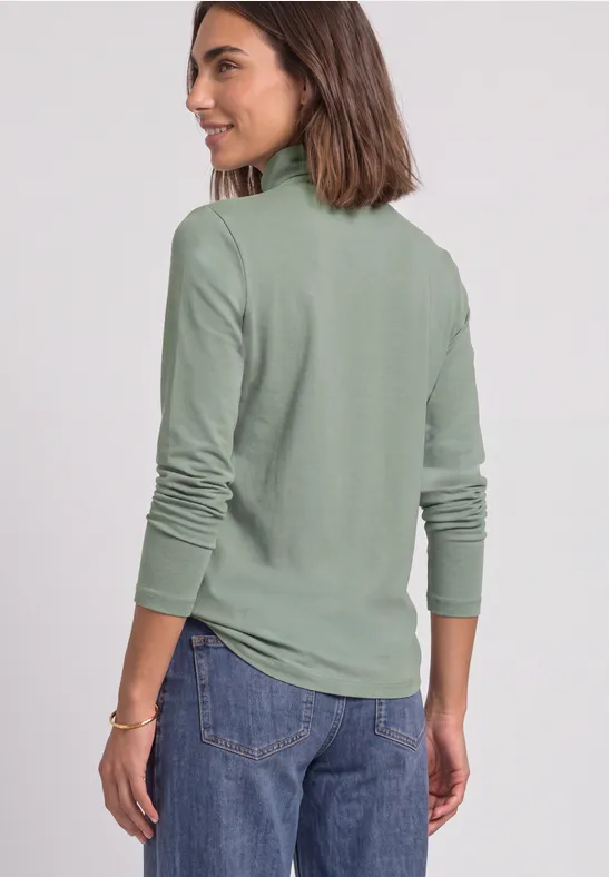 Frau in olivgrüner Rollkragenlongsleeve und Jeans, schaut lächelnd seitlich.