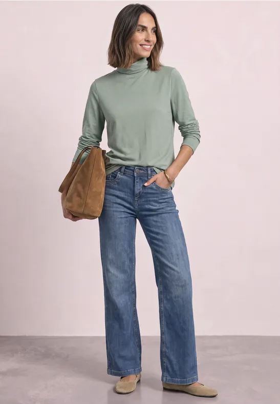 Frau mit grünem Rollkragenpullover, geraden blauen Jeans und brauner Tasche lächelt vor hellem Hintergrund.