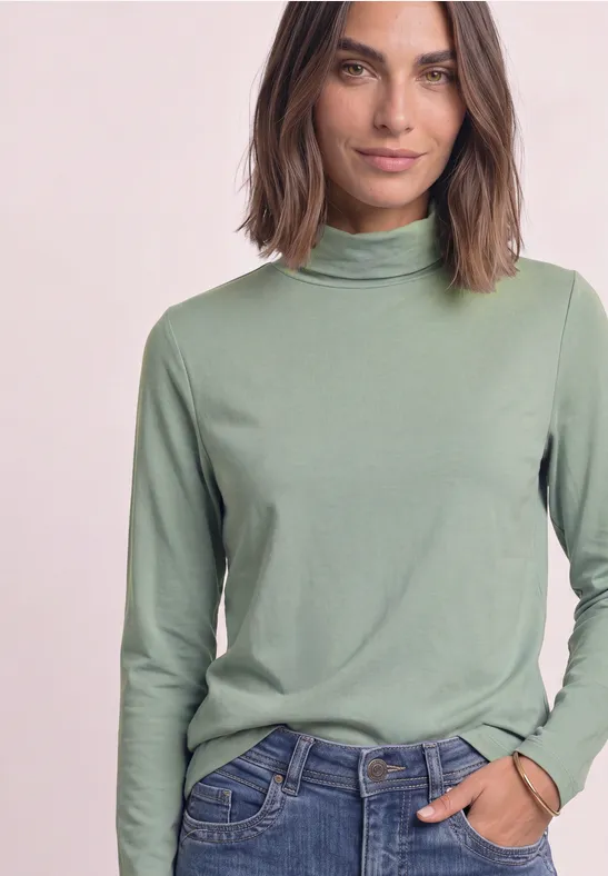 Frau mit schulterlangem Haar trägt grünen Rollkragenpullover und Jeans, eine Hand in der Tasche.