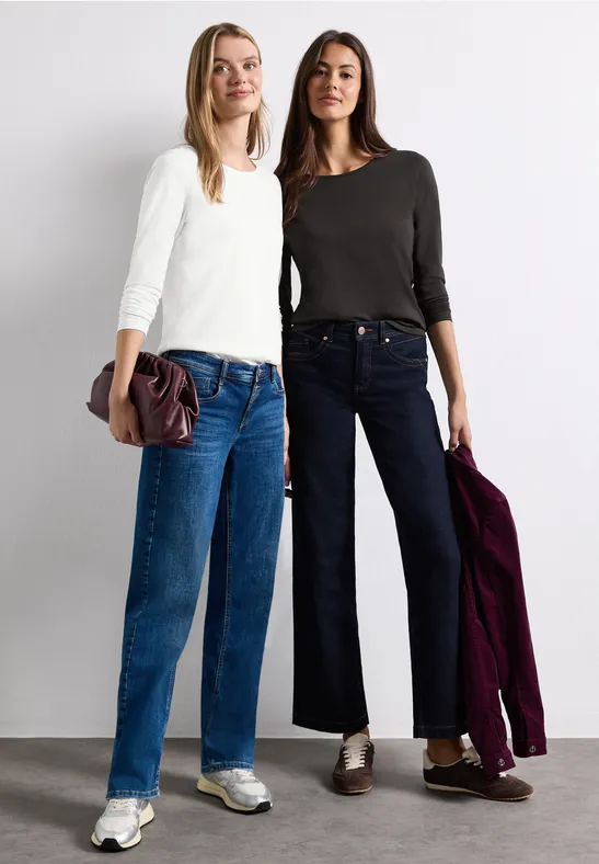 Zwei Frauen posieren in lässiger Alltagskleidung: helles Oberteil und Jeans, dunkler Pullover und weite Hosen.