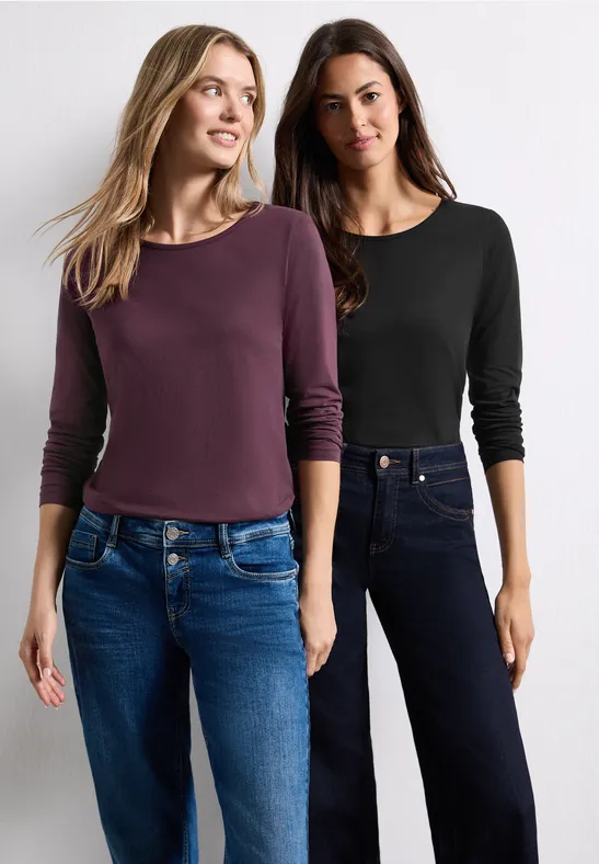 Zwei Frauen in langärmligen Oberteilen und Jeans stehen nebeneinander vor weißem Hintergrund.
