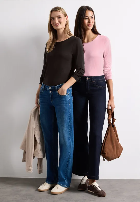 Zwei Frauen in lässiger Mode: Jeans und Pullover, eine hält eine Jacke, die andere eine Tasche.
