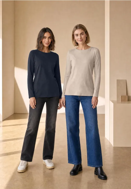 Zwei Frauen posieren in einem hellen Raum, tragen Langarmshirts und weite Jeans.