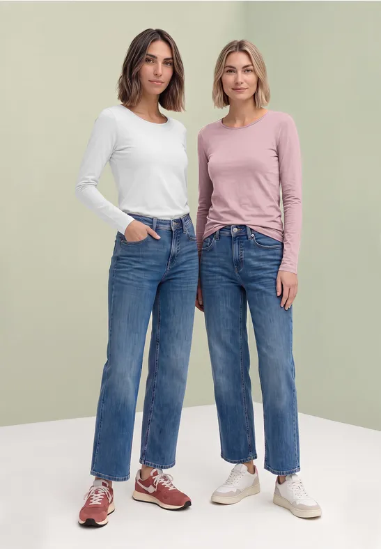 Zwei Frauen in Jeans und langärmligen Shirts posieren vor hellgrünem Hintergrund.