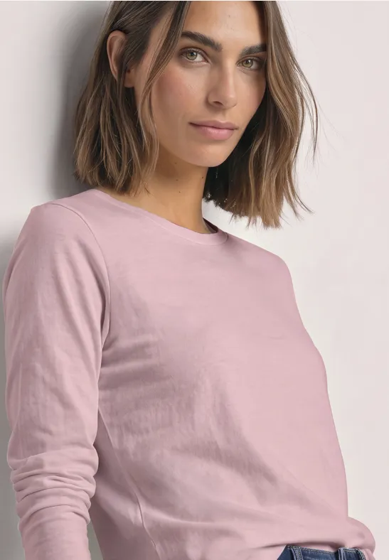Frau mit schulterlangem braunem Haar in rosa Langarmshirt vor hellem Hintergrund.