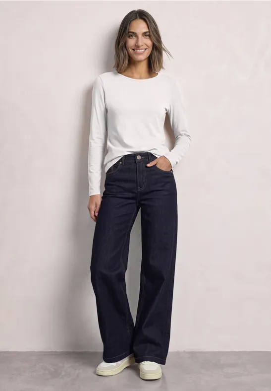 Eine Frau trägt ein weißes Langarmshirt und weite, dunkle Jeans und lächelt vor hellem Hintergrund.