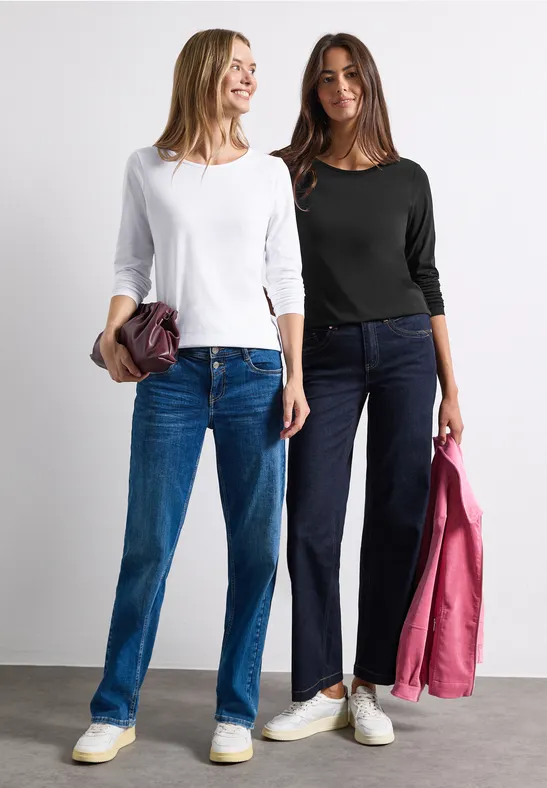 Zwei lächelnde Frauen in Jeans und Langarmshirts, jeweils mit Jacke oder Tasche in der Hand.