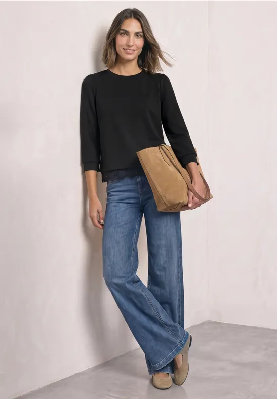 Frau trägt schwarzen Pullover, weite Jeans und hält eine braune Tasche vor heller Wand.