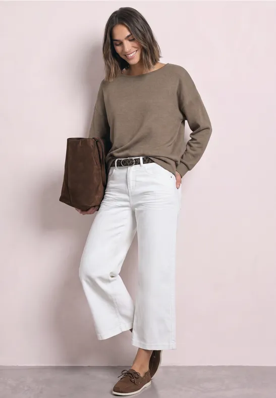 Frau mit taupefarbenem Pullover, weißen Culotte-Jeans und braunen Loafers, trägt braune Wildlederjacke.