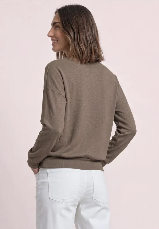 Frau in braunem Pullover und weißen Hosen, seitlich stehend mit Händen in den Taschen vor hellem Hintergrund.