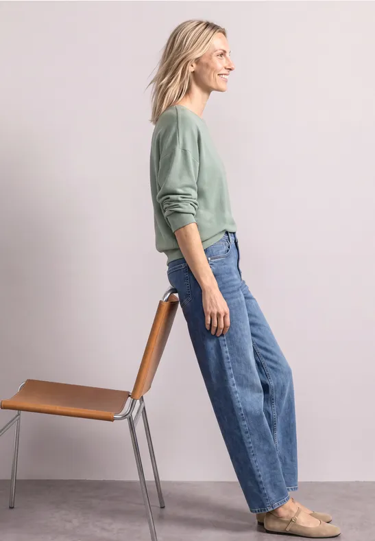 Dame im grünen Pullover lehnt an Stuhl, trägt weite Jeans und flache Schuhe