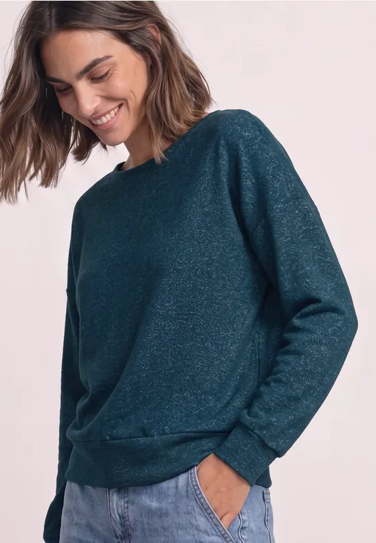 Frau lächelt in einem dunkelgrünen, leicht glitzernden Pullover mit langen Ärmeln und Taschen.