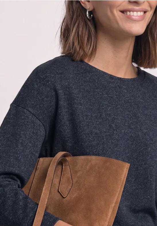 Vrouw in donkergrijze trui lacht en houdt een bruine suède clutch vast.
