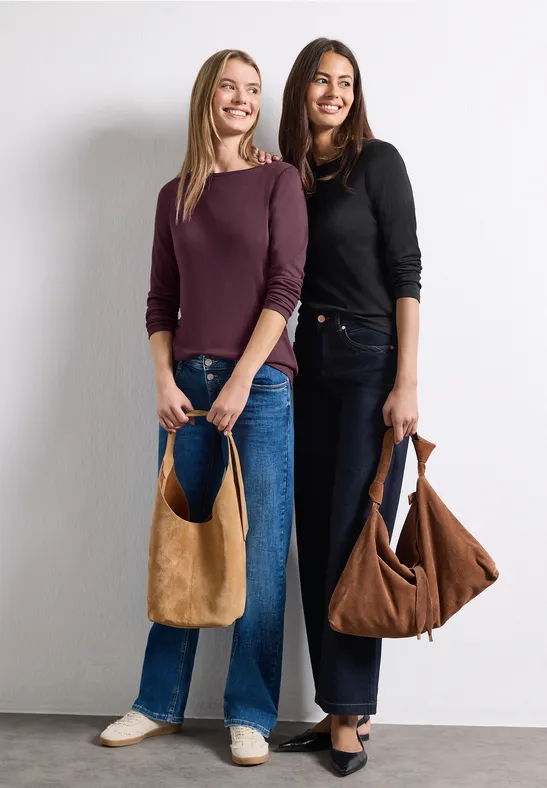 Zwei lächelnde Frauen in Jeans halten braune Taschen vor weißem Hintergrund.