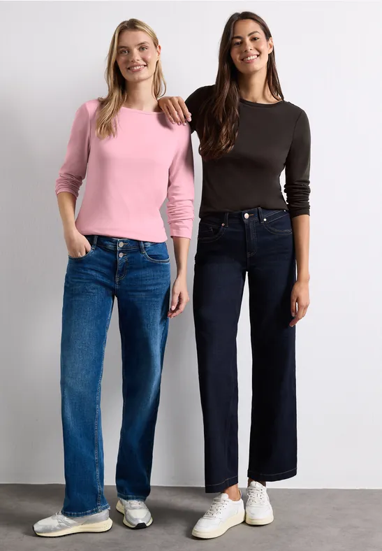 Zwei Frauen in lässigen Langarmshirts und weitem Jeans, lächelnd vor neutralem Hintergrund.