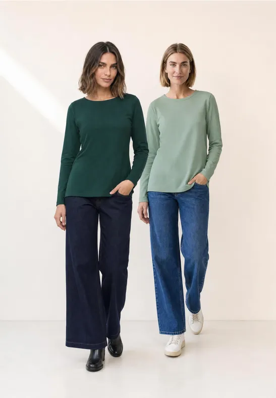 Twee vrouwen in groene truien en jeans staan naast elkaar voor een neutrale achtergrond.