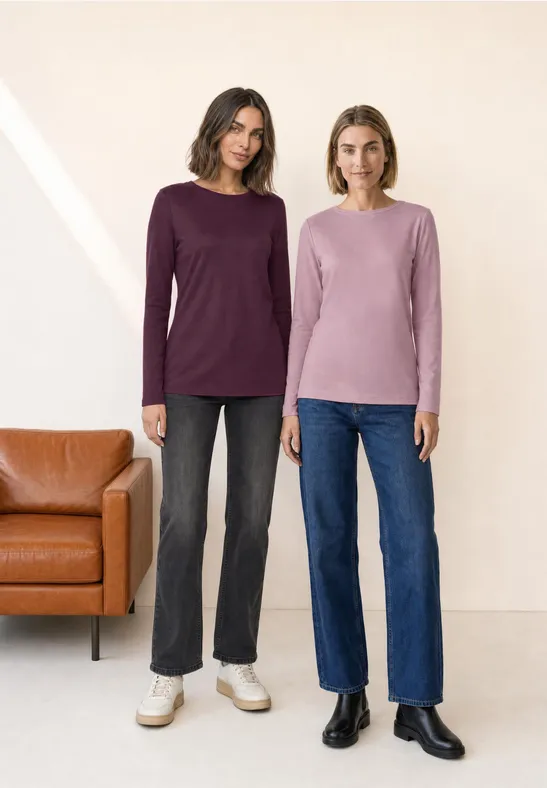 Zwei Frauen in Pullovern und Jeans stehen vor einer hellen Wand neben einem Ledersofa.