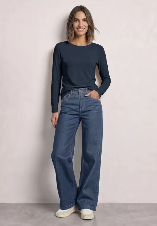 Eine Frau trägt ein langärmeliges dunkelblaues Shirt, weite Jeans und weiße Sneaker vor hellem Hintergrund.