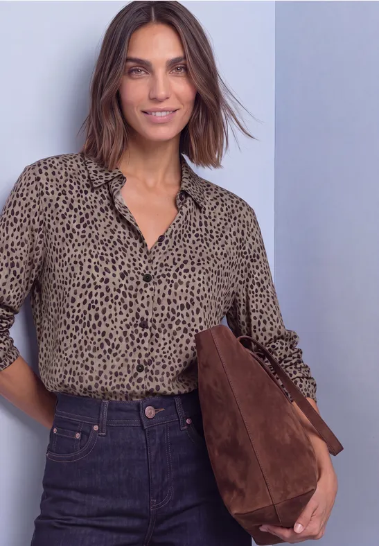 Eine Frau trägt eine beige Bluse mit schwarzen Punkten, dunkle Jeans und eine braune Wildledertasche.