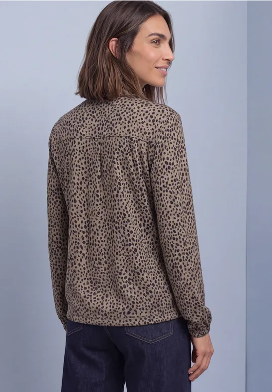Frau von der Seite, trägt beige-braune Bluse mit schwarzem Leoprint und dunkler Hose vor schlichtem Hintergrund.