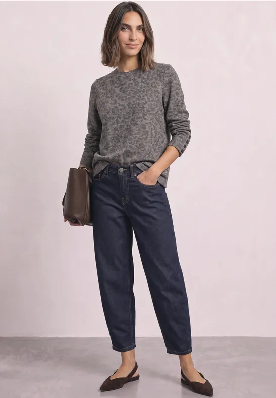 Frau trägt grauen Pullover, dunkelblaue Jeans, schwarze Ballerinas, hält braune Tasche vor hellem Hintergrund.