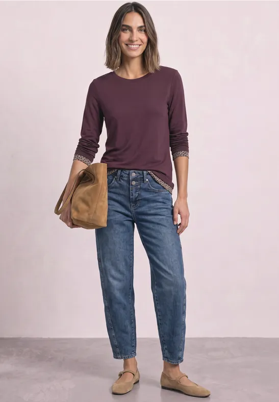 Frau in Jeans, langärmeligem Oberteil, Slippern und Tasche vor hellem Hintergrund.