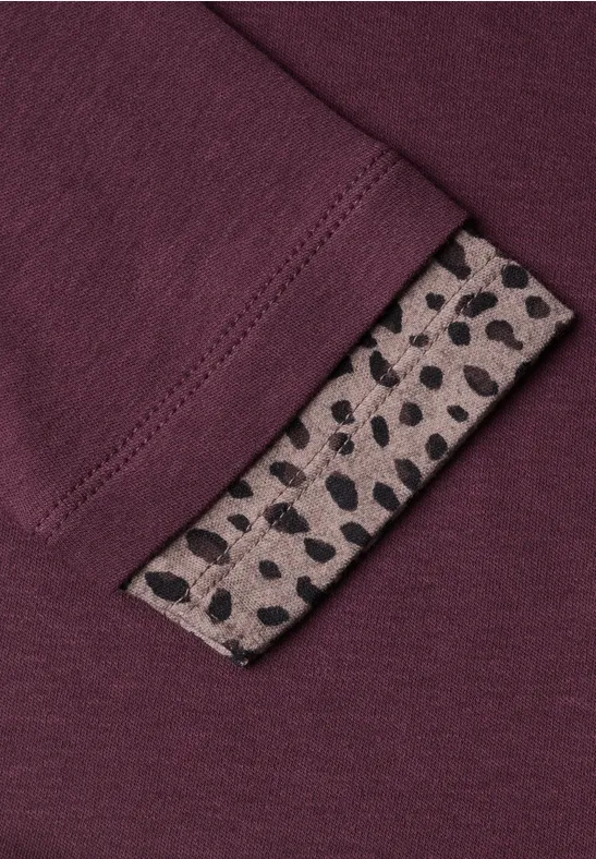 Bordeauxrotes Textil mit einem beigen Label, das schwarze, unregelmäßige Punkte zeigt.