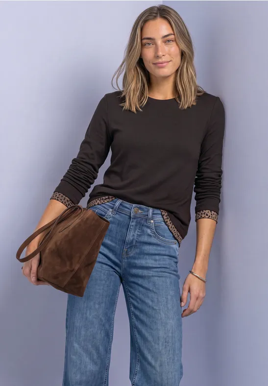 Eine Frau trägt schwarzen Pullover, blaue Jeans und hält eine braune Tasche vor violettem Hintergrund.
