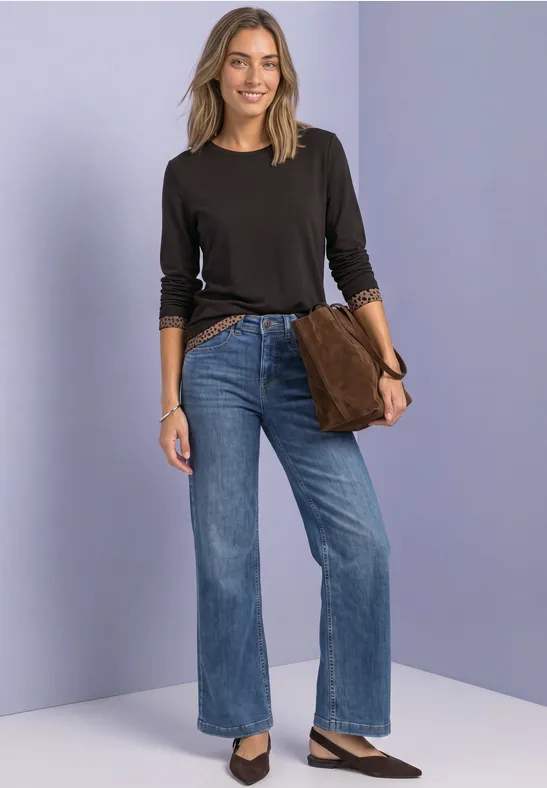 Frau in schwarzem Langarmshirt, weitem Jeans, schwarzen Sandaletten und brauner Clutch vor pastellblauem Hintergrund.