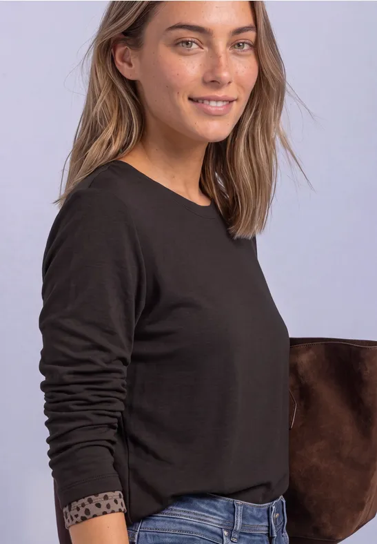 Junge Frau trägt ein schlichtes schwarzes Langarmshirt mit dezenten Punkten am Ärmelsaum vor hellem Hintergrund.