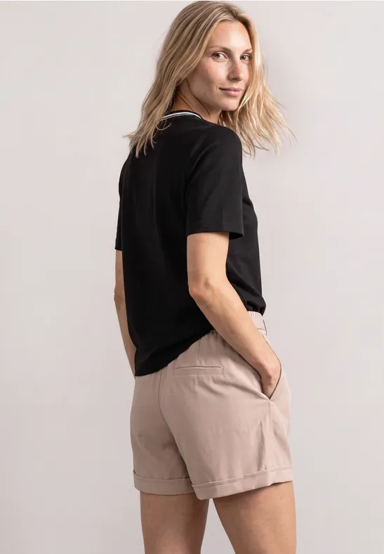 Frau mit blondem Haar in schwarzem T-Shirt und beigefarbenen Shorts steht seitlich, eine Hand in der Tasche.