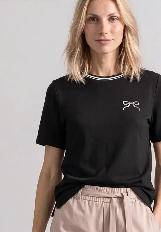 Eine Frau trägt ein schwarzes T-Shirt mit kleinem Schleifendruck und beigen Hosen vor neutralem Hintergrund.