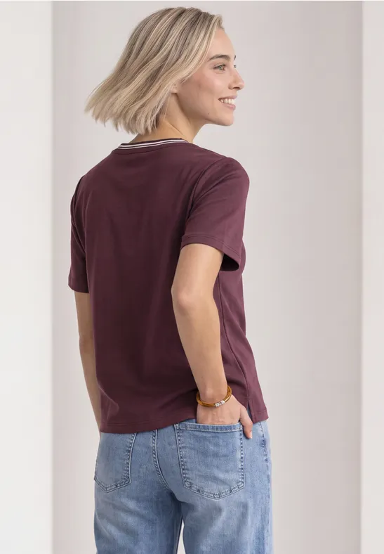 Eine Frau mit kurzen blonden Haaren trägt ein weinrotes T-Shirt und eine blaue Jeans, von hinten zu sehen.