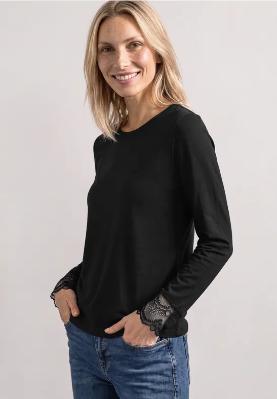 Uśmiechnięta blondynka ma na sobie czarną bluzkę z długimi rękawami i koronkowymi mankietami oraz jeansy.
