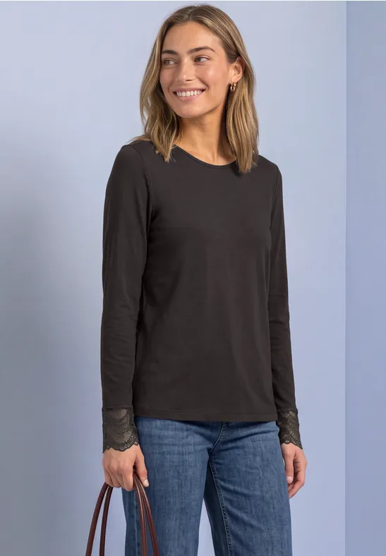 Junge Frau lächelt, trägt schwarz langärmliges Shirt, blaue Jeans und hält eine braune Tasche.