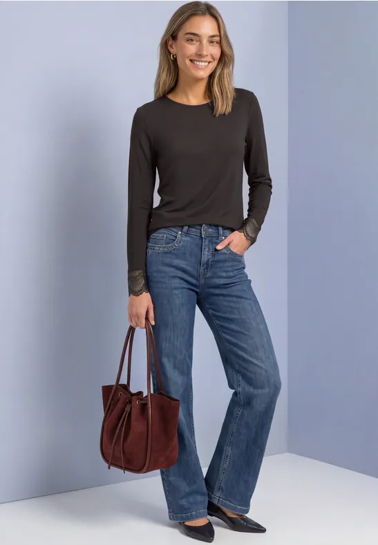 Junge Frau in schwarzem Langarmshirt und weiten Jeans hält eine braune Ledertasche vor neutralem Hintergrund.