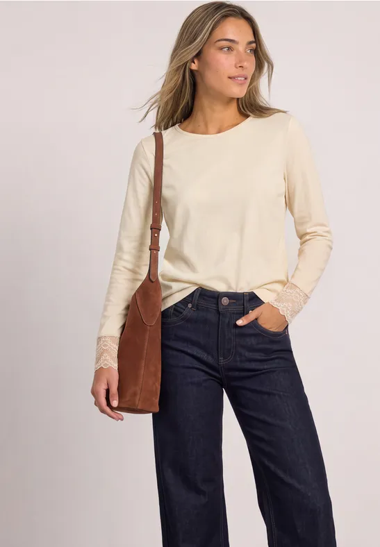 Frau mit cremefarbenem Langarmshirt mit Spitze, dunkelblauer Jeans und brauner Umhängetasche.