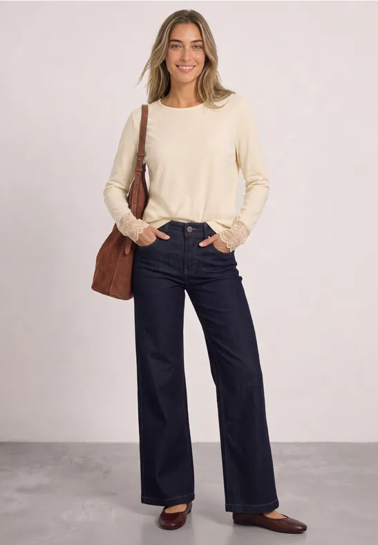 Eine Frau in einem beigen Pullover, weiten dunkelblauen Jeans und braunen Schuhen mit Schultertasche.