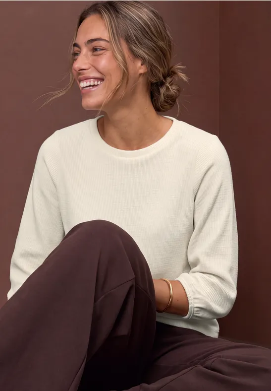 Eine junge Frau sitzt lächelnd, trägt beigen Pullover und braune Hose vor braunem Hintergrund.