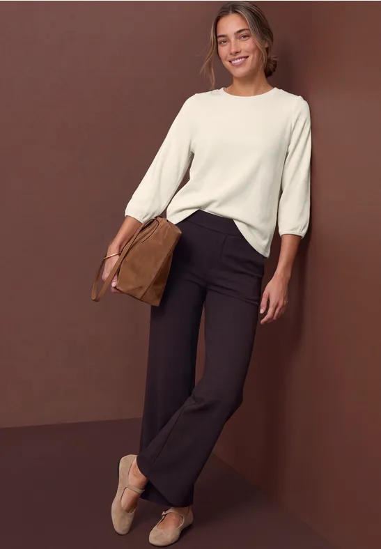 Frau lehnt an einer braunen Wand, trägt weiße Bluse, schwarze Hose, beigefarbene Slingback und braune Tasche.